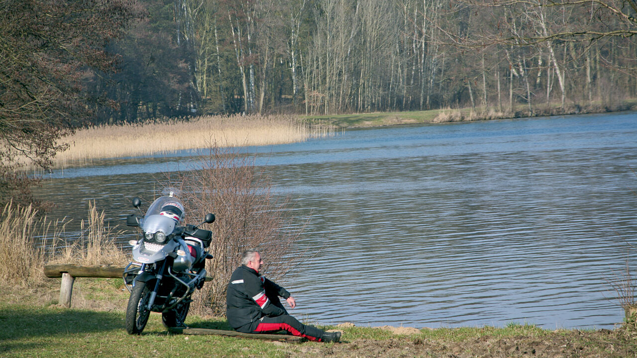 Motorradfahrer sitzt am Ufer neben seiner Maschine, vor ihm ein See mit Schilf